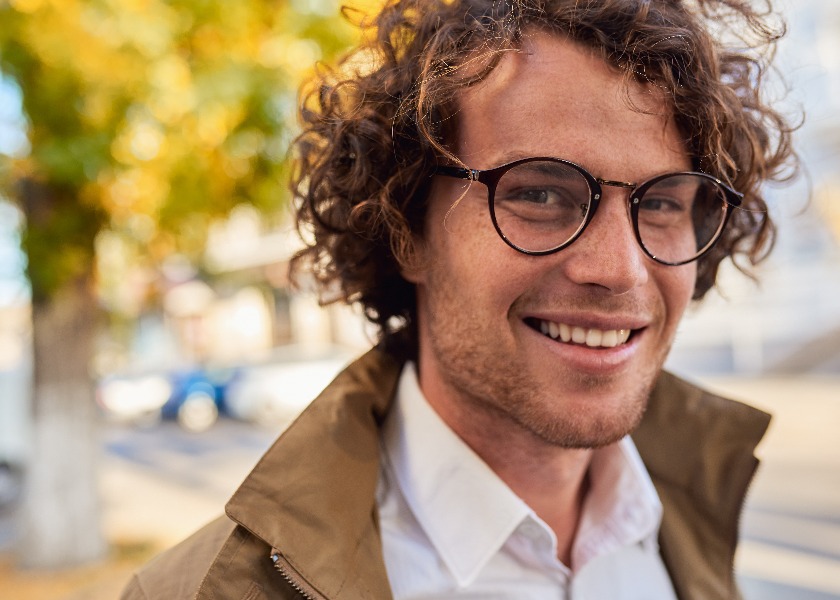 Closeup horizontal portrait of young happy business man with glasses smiling and posing outdoors. Male student in autumn street. Smart guy in casual wears spectacles with curly hair walking on street